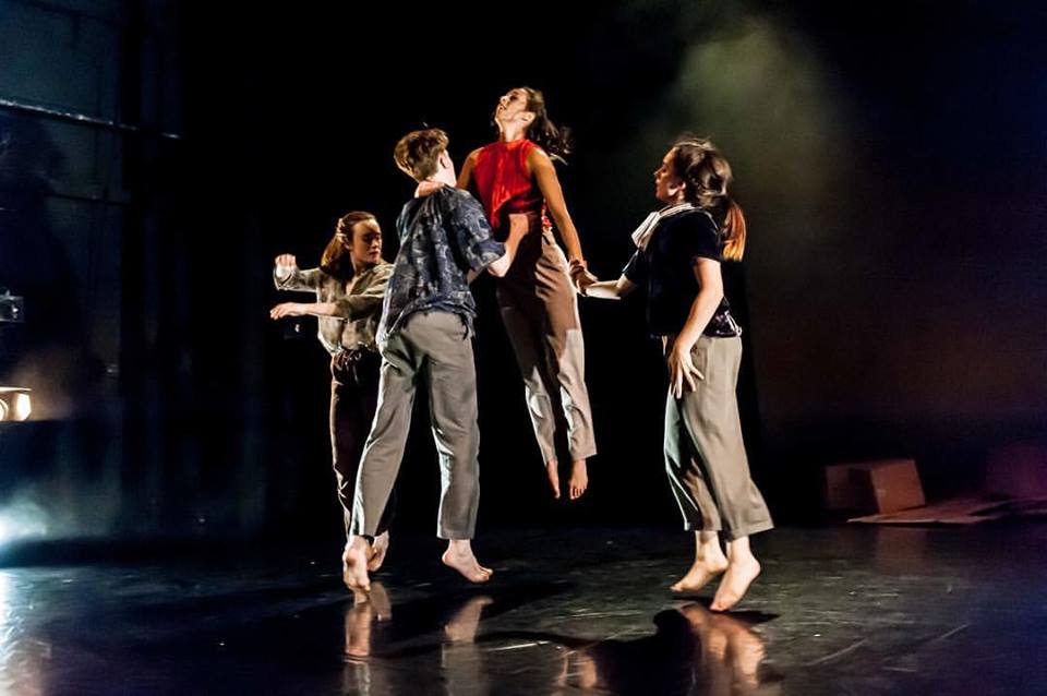 Four young contemporary dancers, one being lifted into the air. Black backdrop with stage lights.