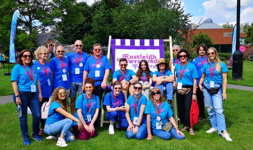 A large group of staff and volunteers wearing blue Eastleigh Unwrapped T-Shirts and red lanyards face the camera. They are smiling and some sit on a giant branded deckchair.